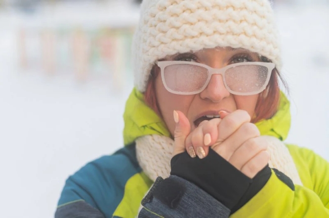 Portrait of a woman in glasses covered with hoarfrost. The girl is freezing and forgot gloves in very cold weather and blows on her bare hands